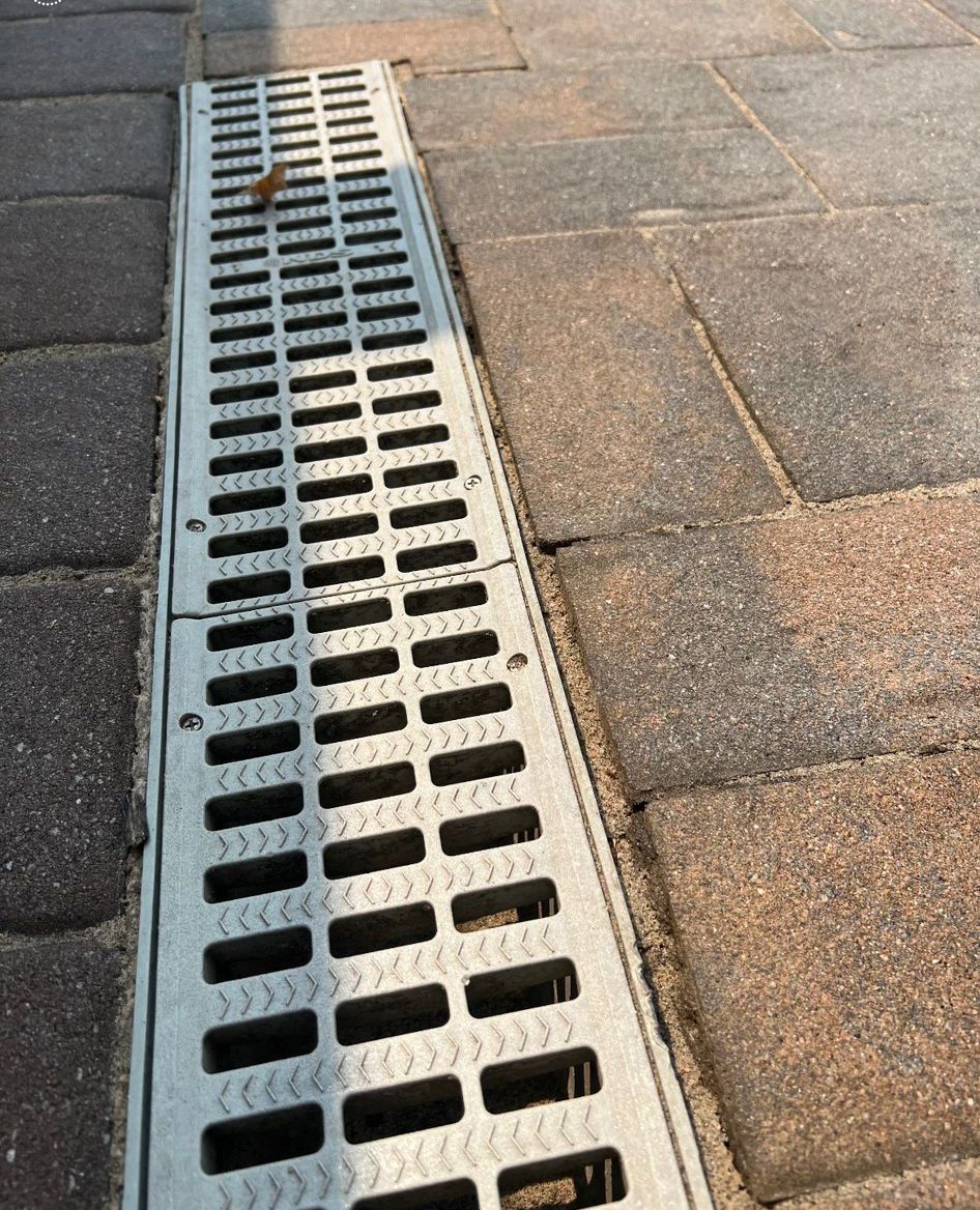 Metal drainage grate beside a stone-paved sidewalk, viewed at an angle