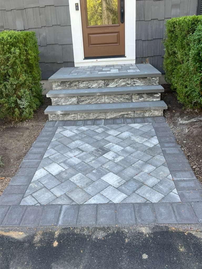 Front steps and a brown door above a decorative stone-paver walkway between hedges.