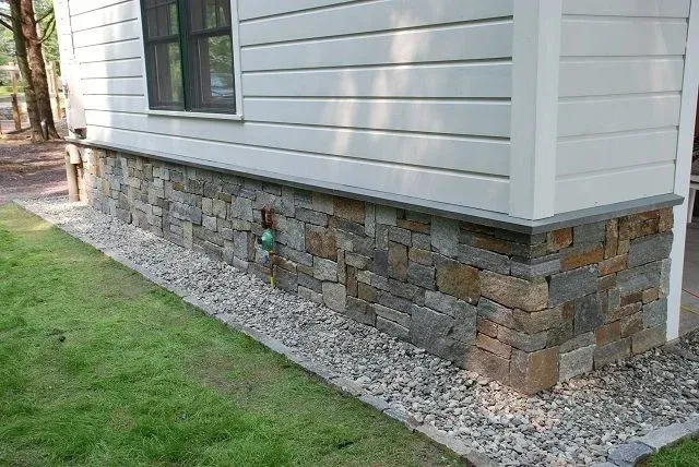 House exterior with gray siding and a stone veneer foundation beside gravel and grass