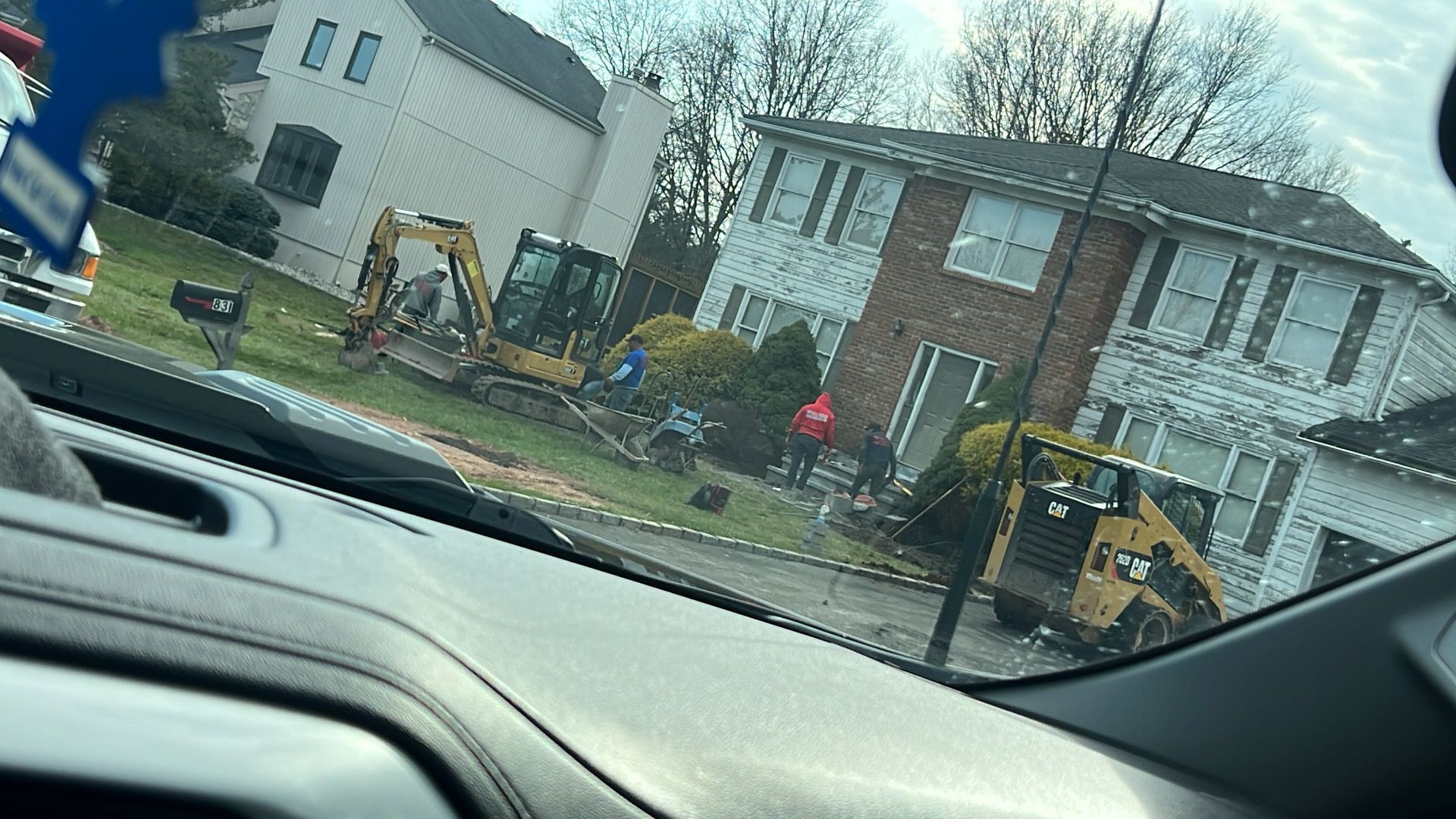 Small yellow excavator in a residential yard beside a house, viewed from a car windshield.