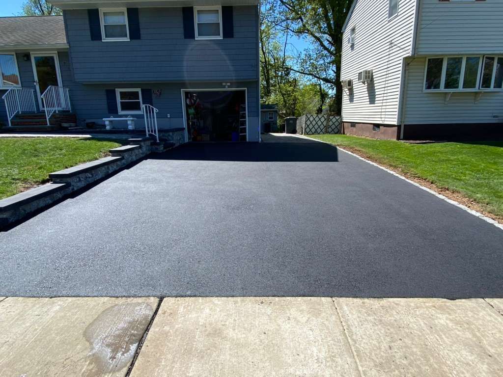 Fresh black asphalt driveway beside a two-story house and lawn on a sunny day