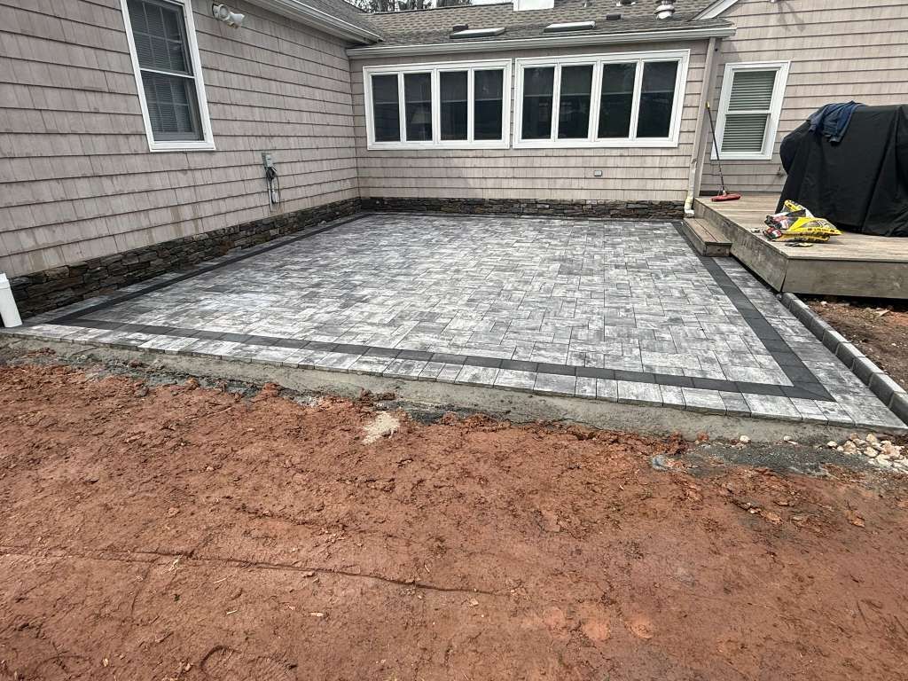 New stone patio beside a house, bordered by gray pavers and a dirt foreground