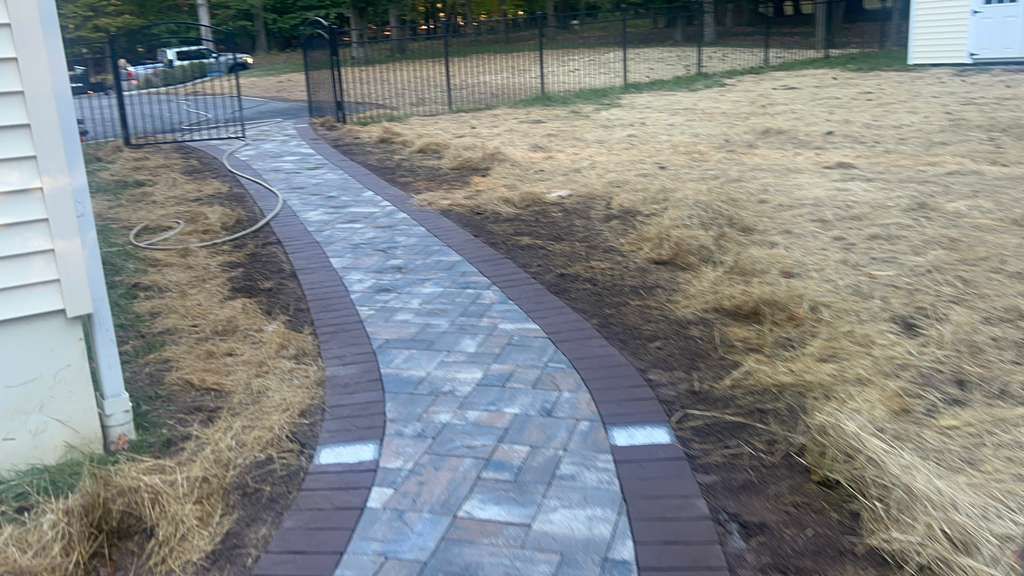 Curved brick walkway through a dry yard beside a house, leading to a fenced gate.