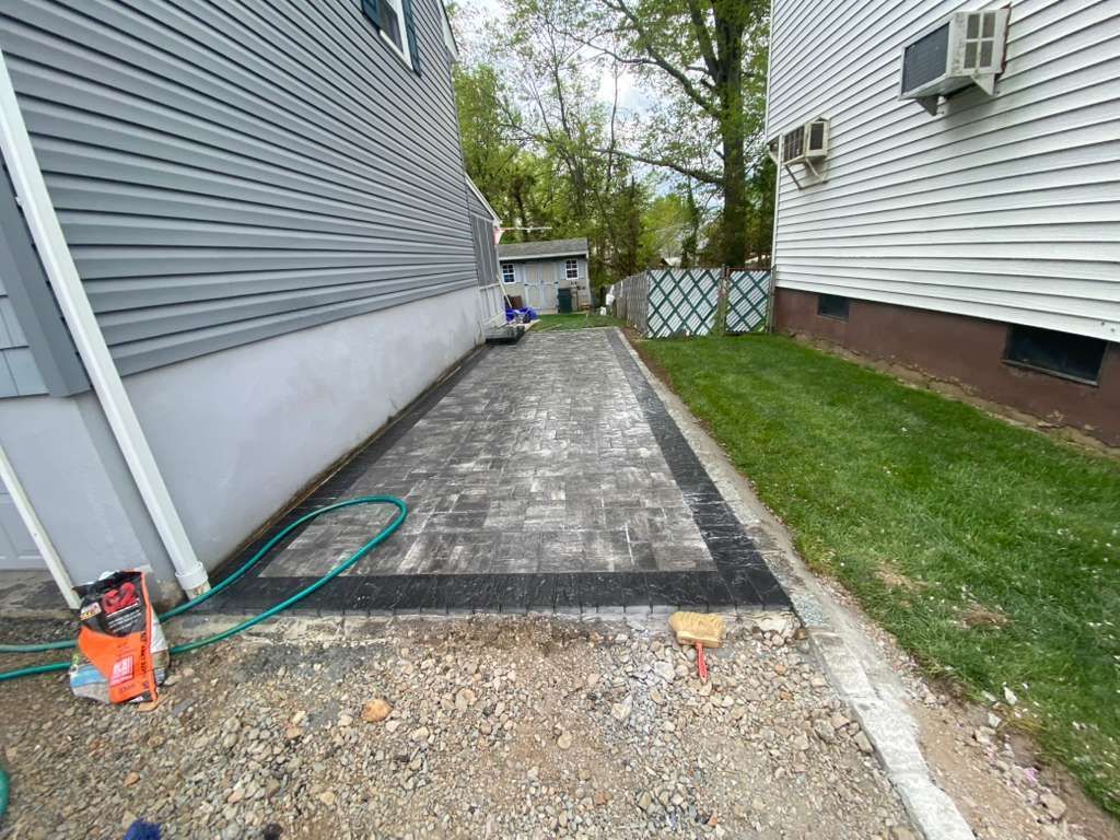 Narrow concrete walkway between houses with a garden hose and orange bag near a gravel edge