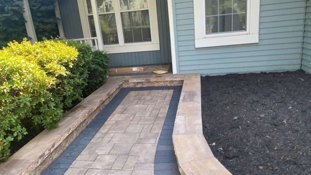 Brick walkway leading to a front door beside a light blue house and black mulch bed