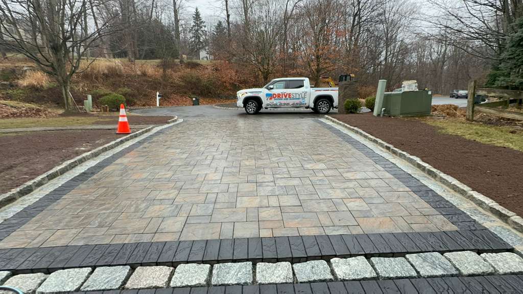 Paved driveway under construction, with a work truck and orange cone at the end.