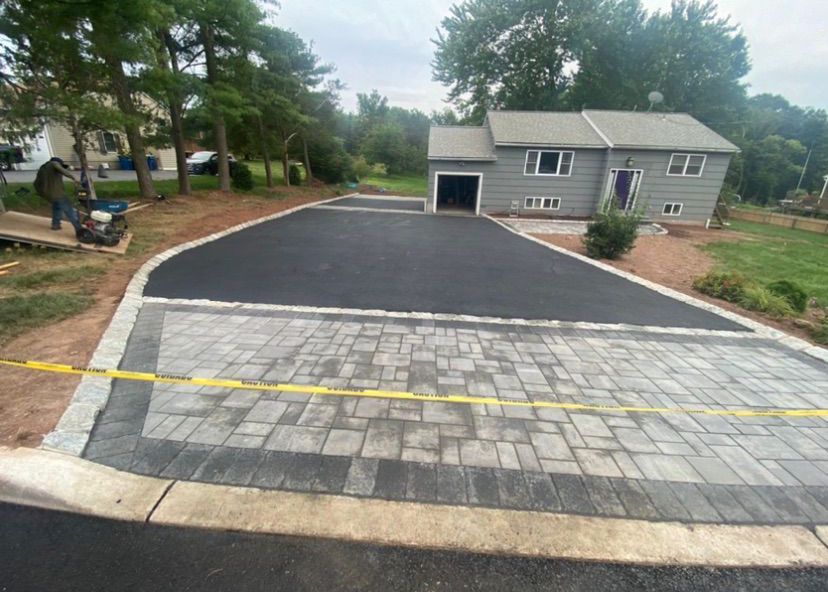 Freshly paved driveway with a gray paver apron leading to a house and garage, bordered by trees and lawn.