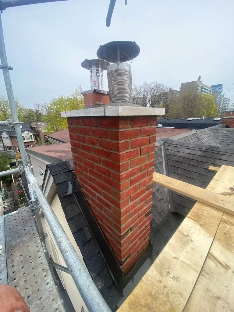 Brick chimney on a rooftop with metal vent cap, surrounded by scaffolding and roofing materials