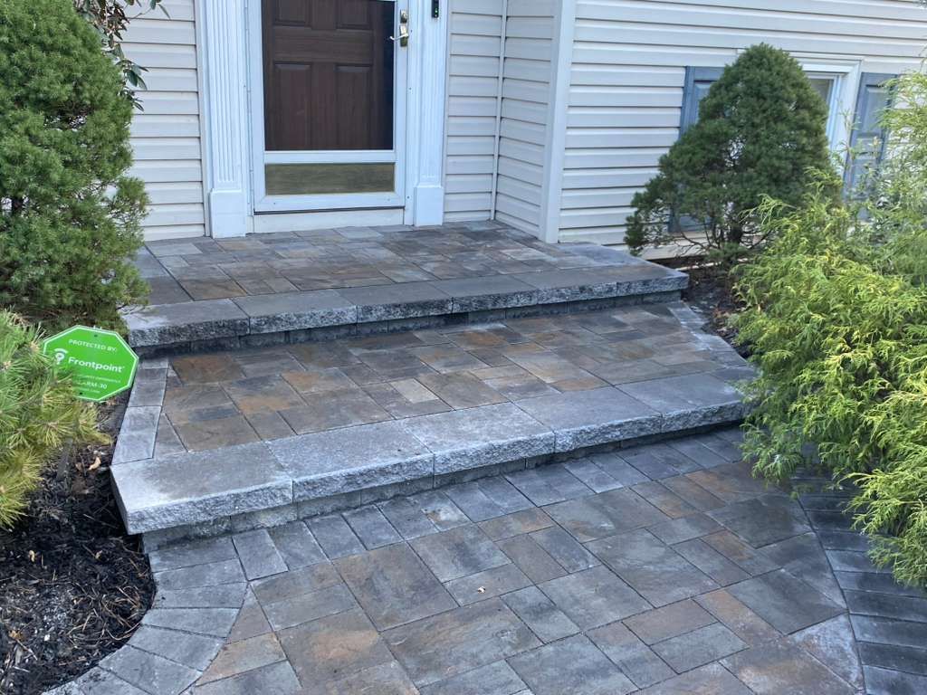 Front entry with stone steps, tiled landing, and white siding, bordered by shrubs.