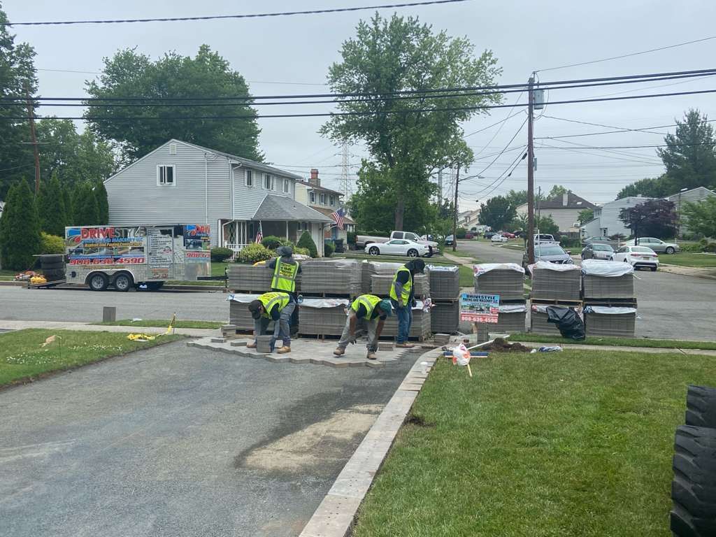 Workers in neon vests paving a driveway at a residential street, with roadwork equipment nearby.