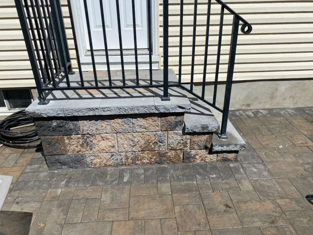 Front stoop with stone steps, black metal railing, and brick paver walkway by a house exterior