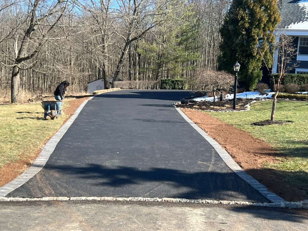 Freshly paved driveway bordered by mulch and trees, with a person working on the left side.