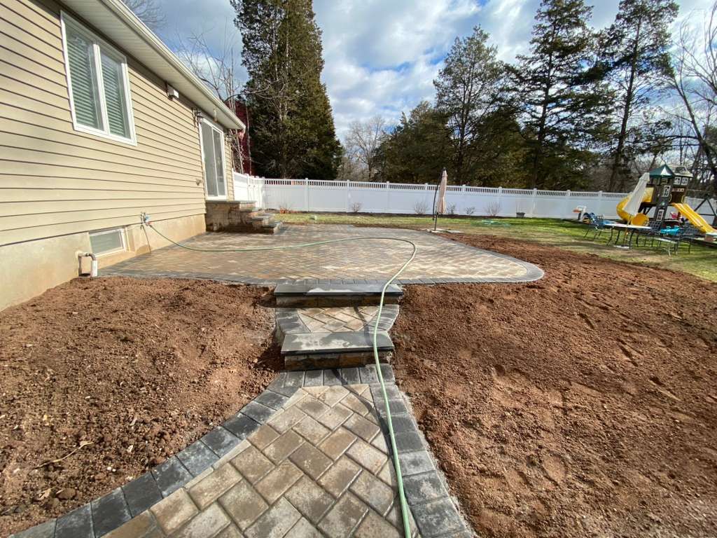 Backyard patio under construction beside a house, with stone steps and a white fence in the background