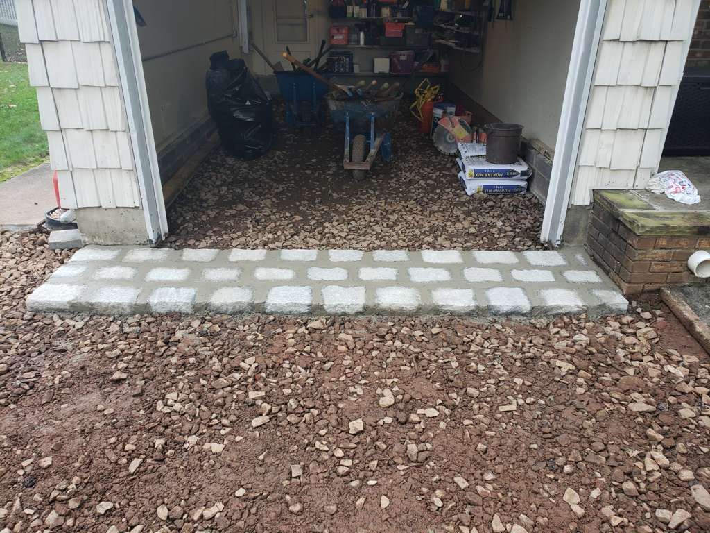 Open garage with tools and storage, stone paver threshold, and gravel driveway outside.