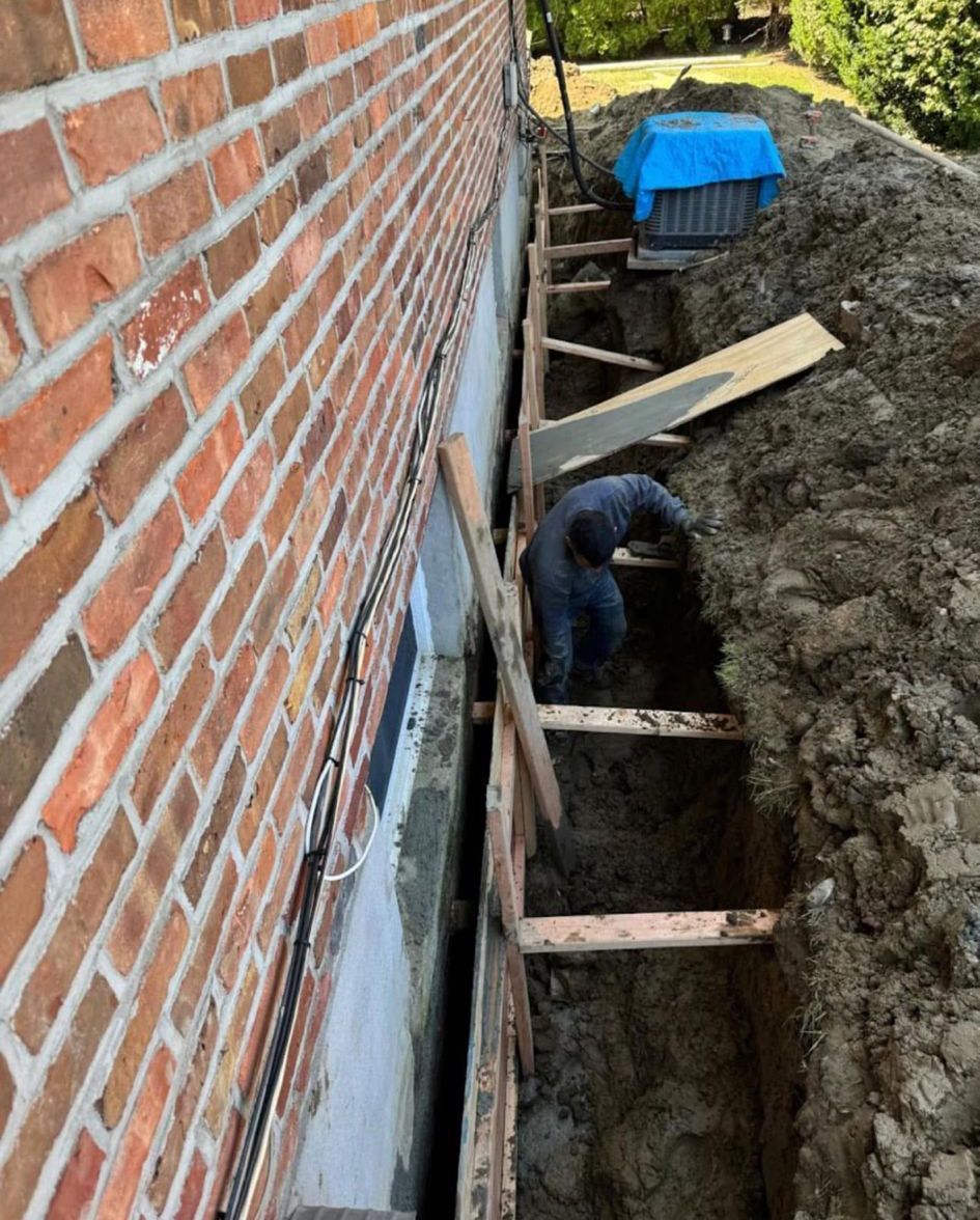 Brick house wall beside a deep trench with wooden supports and a worker digging near a blue bin