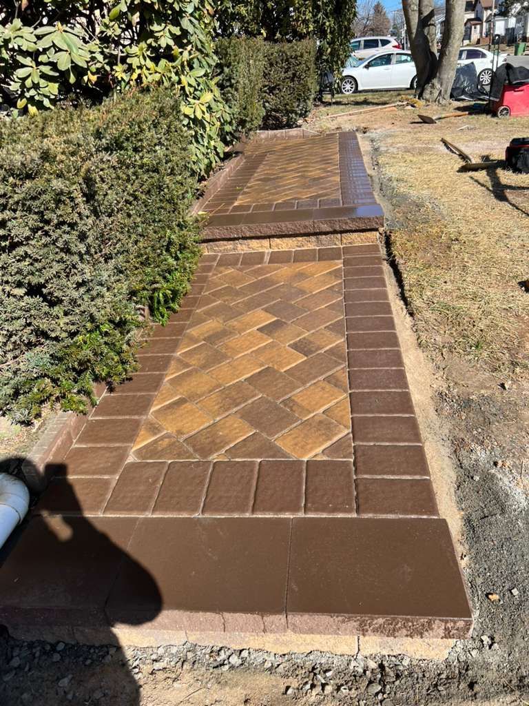 Brick walkway beside hedges and a dry grassy path, leading toward parked cars.