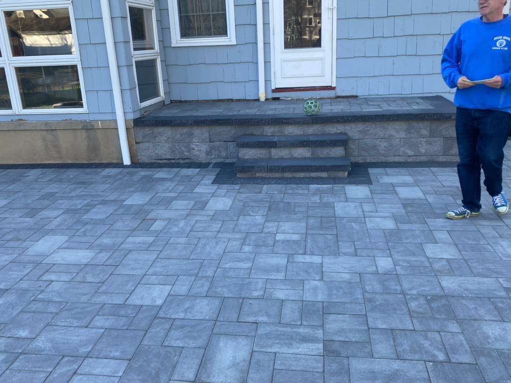 Person in a blue shirt standing on a gray paver patio in front of a house entrance.