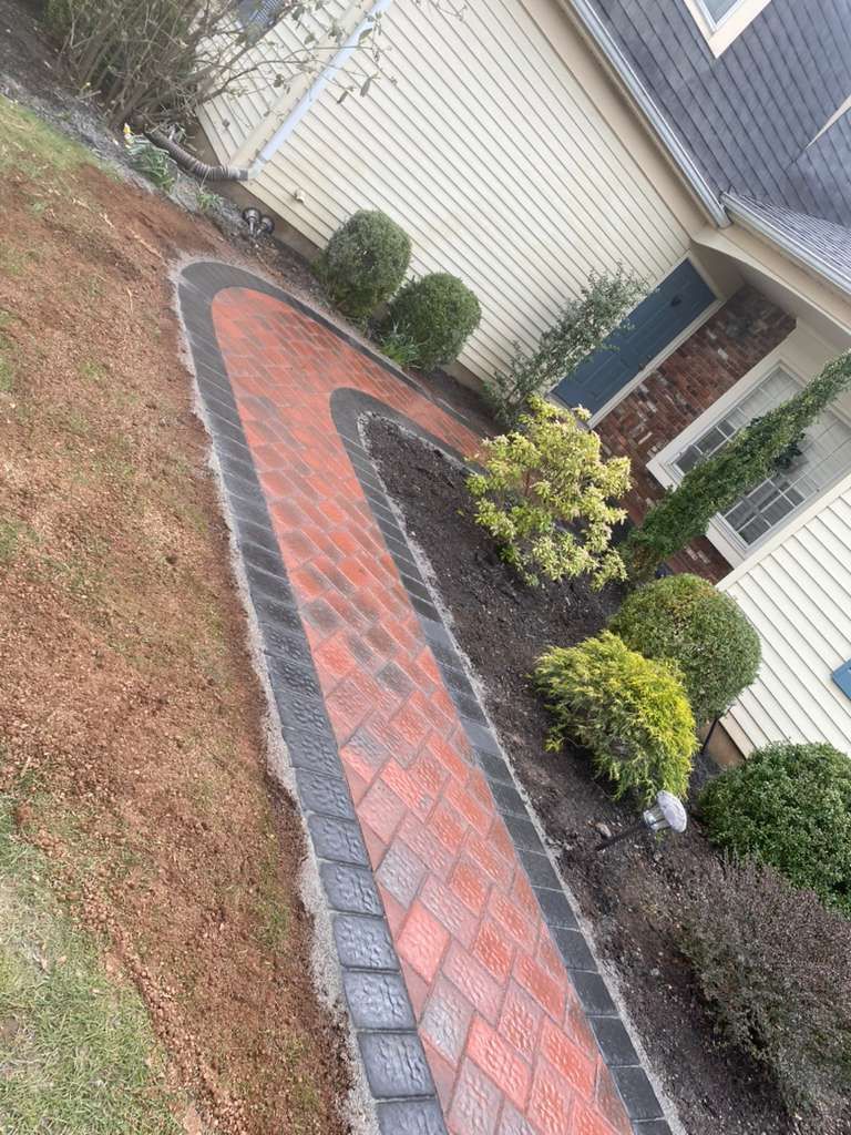 Curved red brick walkway leading to a house entry, bordered by gray pavers, mulch, and shrubs.