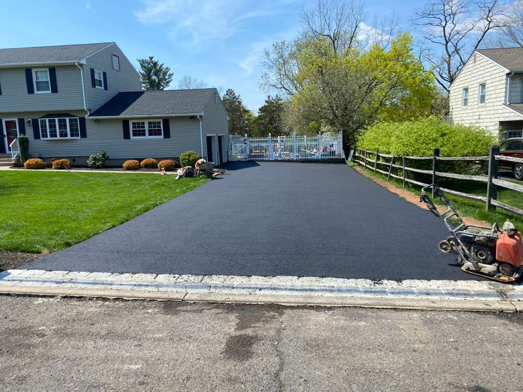 New blacktop driveway between suburban houses, with lawn and white fence at the end