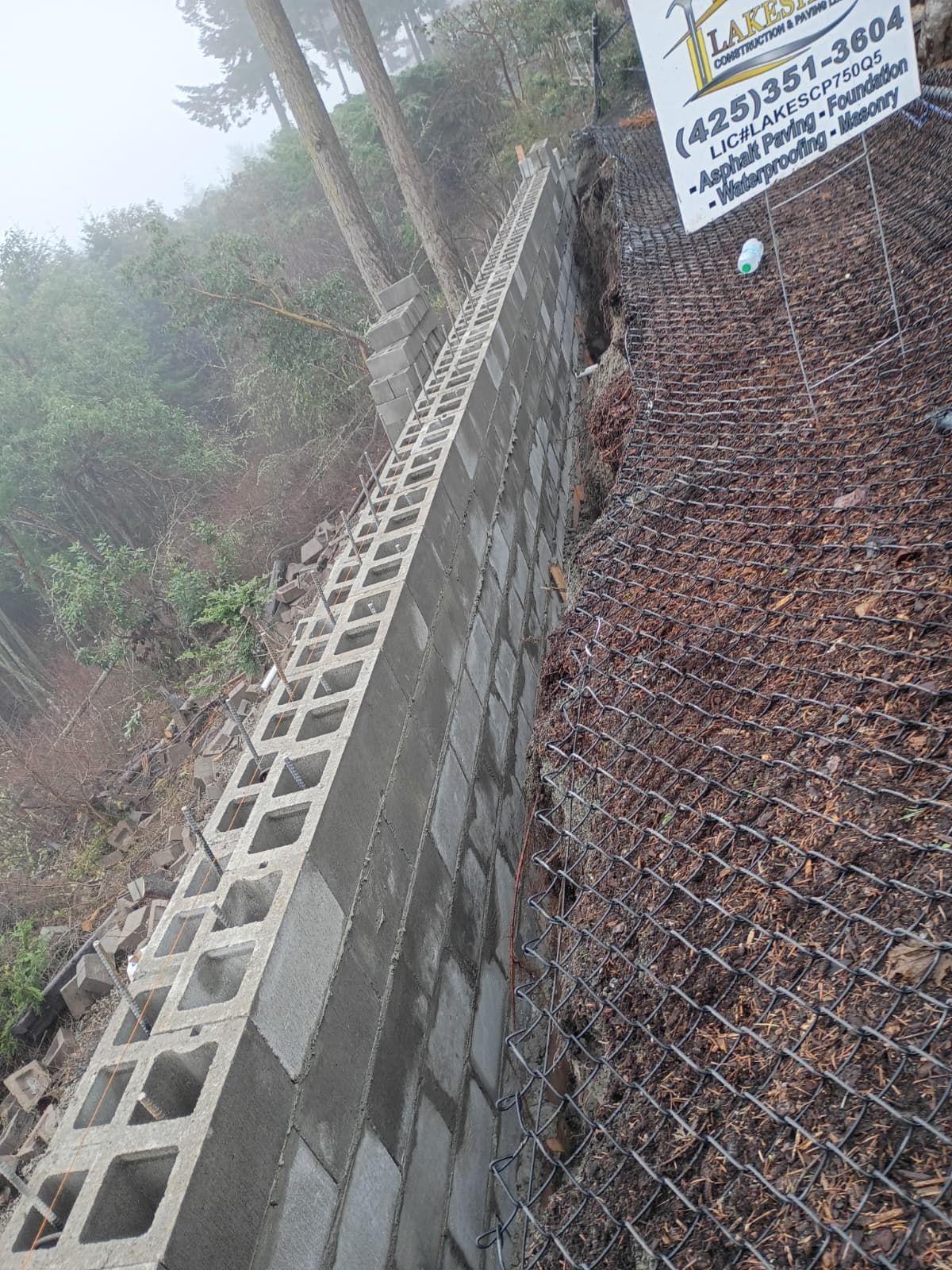Tall concrete retaining wall with cinder blocks along a wooded hillside, with a sign on top.