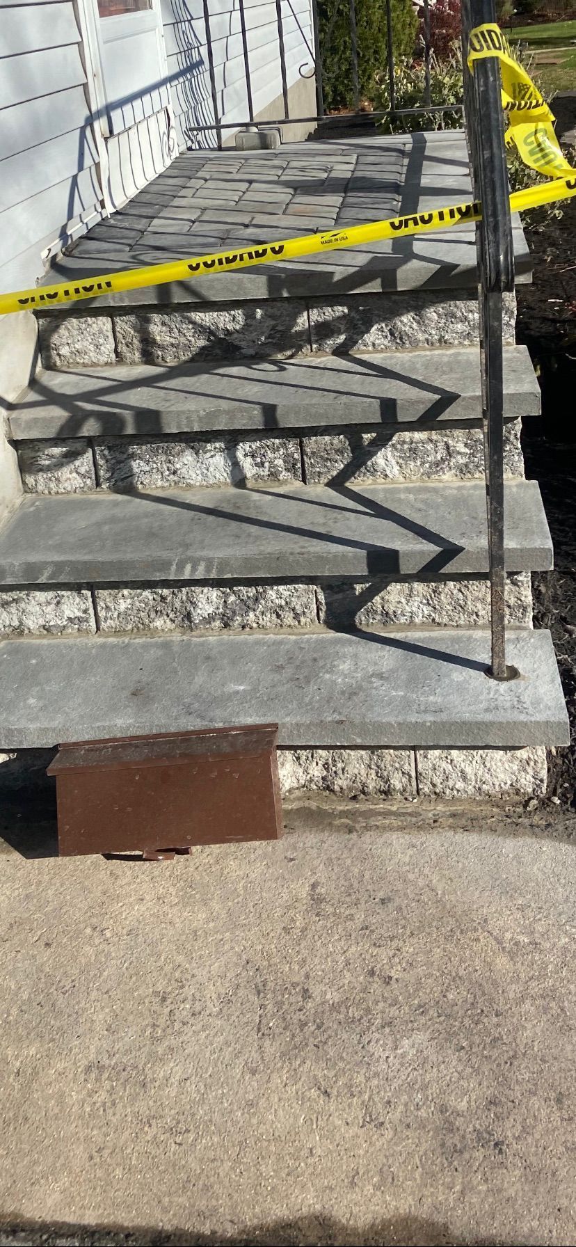 Outdoor stairs blocked by yellow caution tape and a handrail, with a brick on the bottom step.