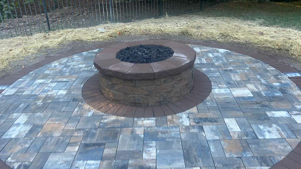 Circular stone fire pit in the center of a paved outdoor patio, surrounded by dry grass.