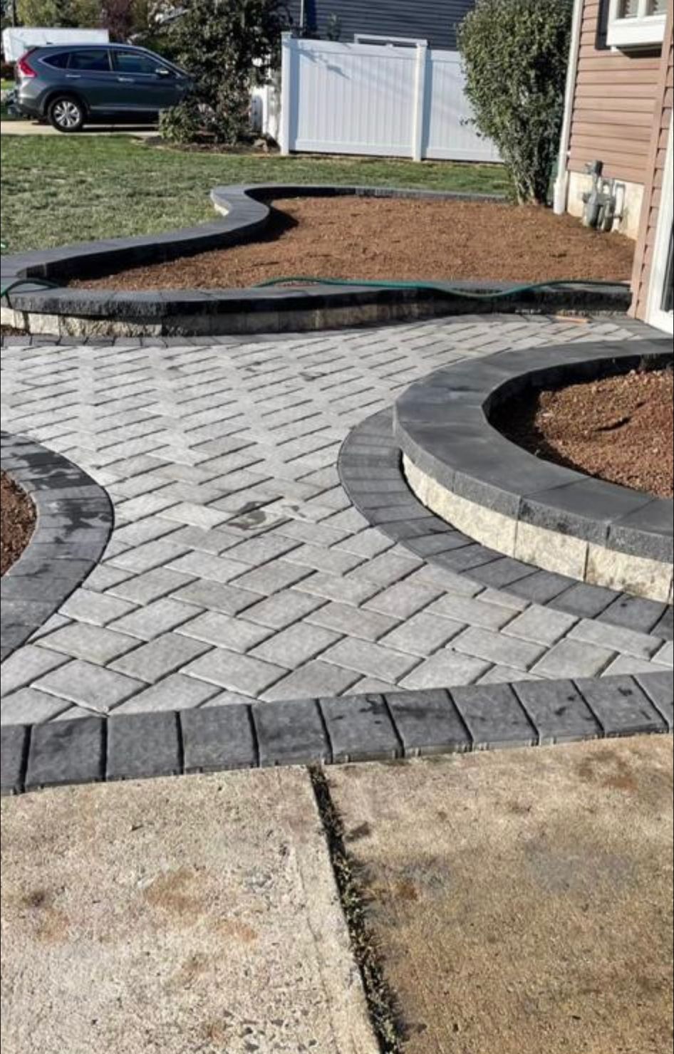 Curved brick walkway with raised mulch beds beside a house and driveway, leading to a white gate.