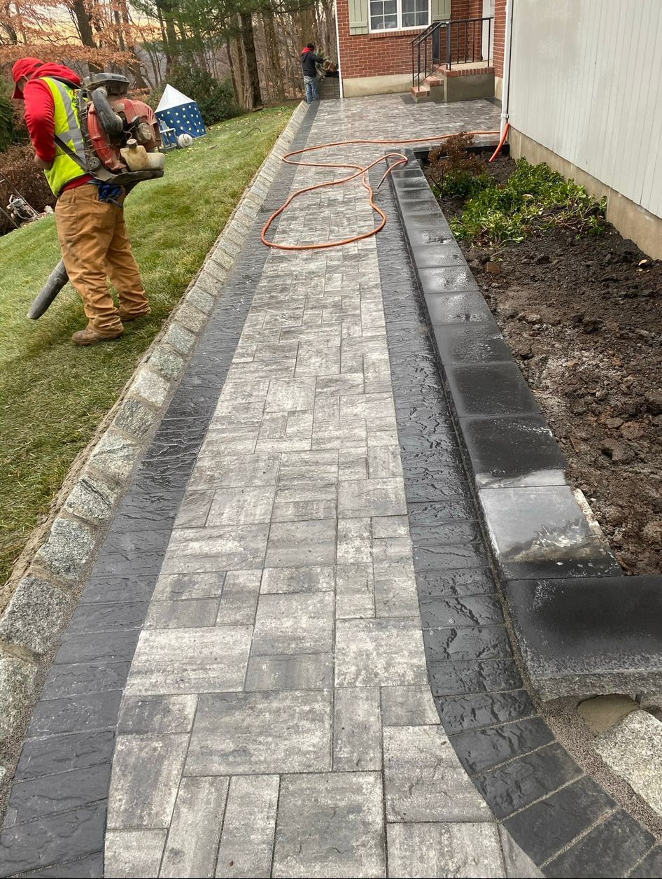 Workers installing gray pavers along a curved walkway beside a black retaining wall and brick house.