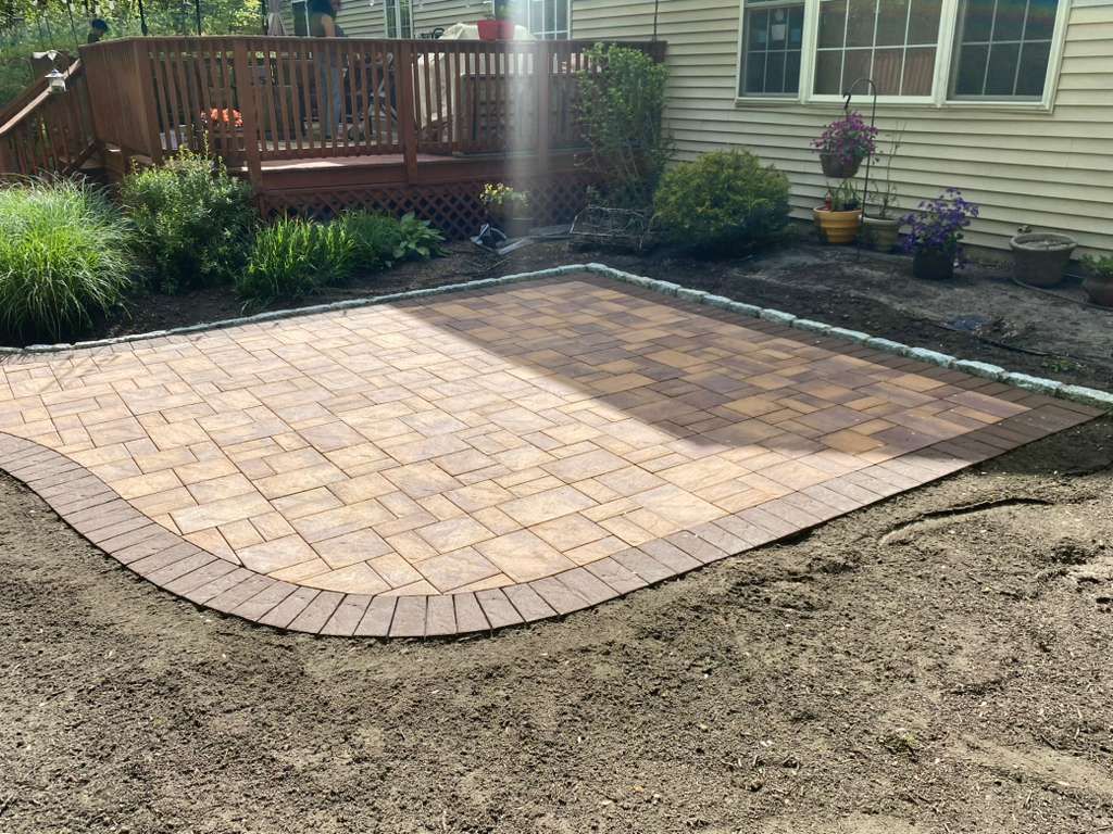 Backyard patio with newly laid brick pavers and curved edging beside a house and deck.