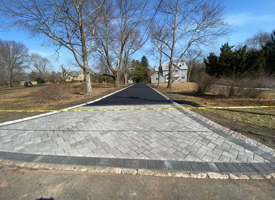 New paved path crossing with yellow caution tape, trees, and houses under a clear blue sky