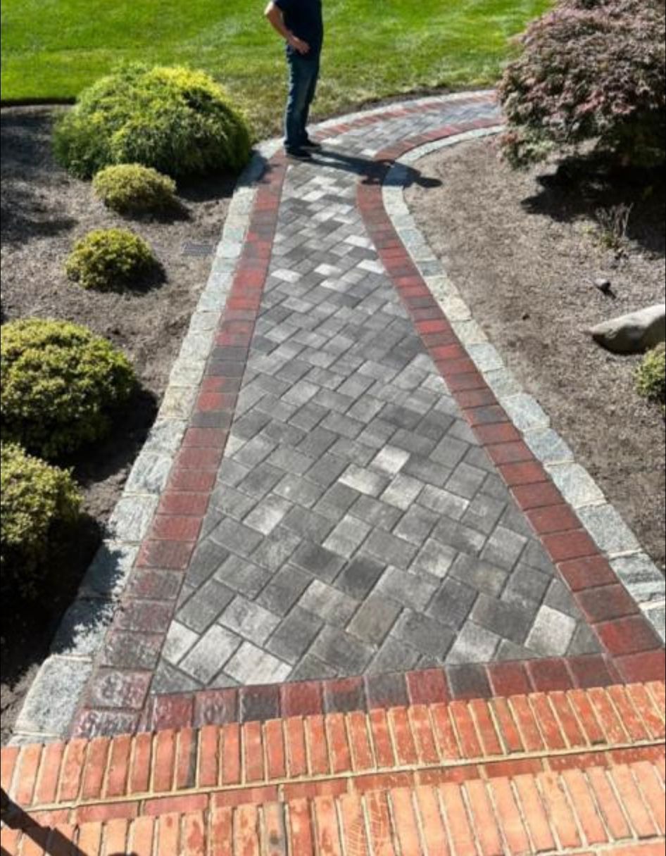 Brick-and-gray paver walkway through a landscaped yard, with a person standing at the far end.