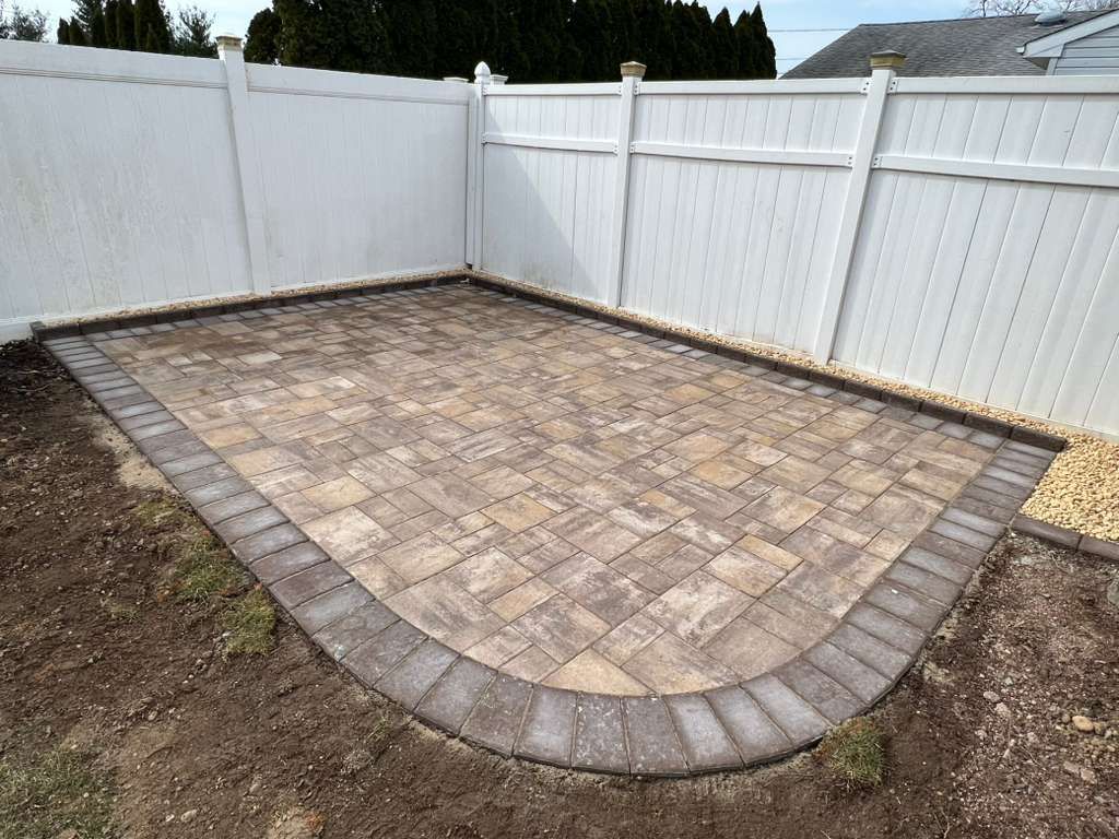 Paver patio bordered by gray stones in a fenced backyard, with bare soil around the edges.