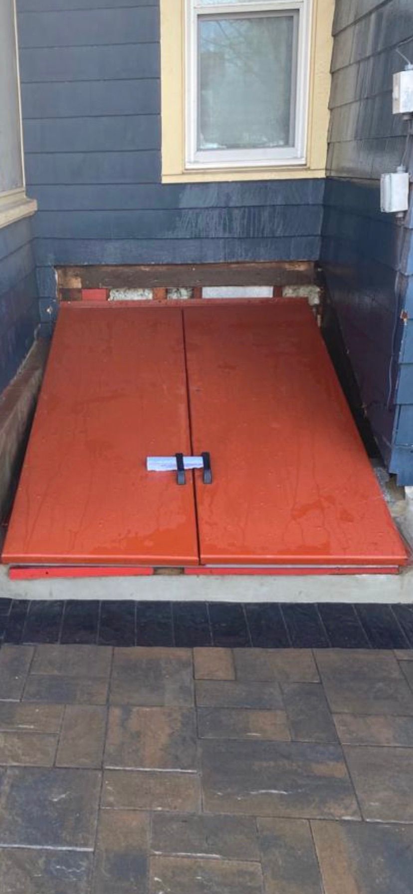 Red basement hatch doors set in a brick patio below a window.