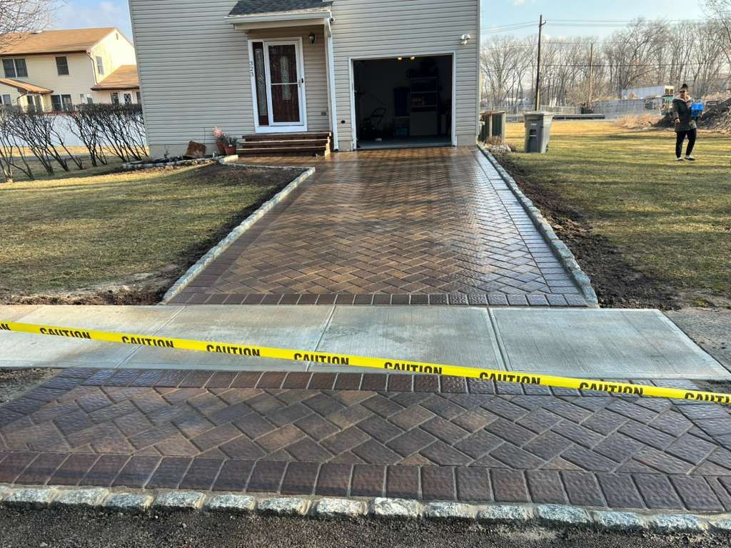 Taped-off brick driveway leading to an open garage and house, with a person standing in the yard.