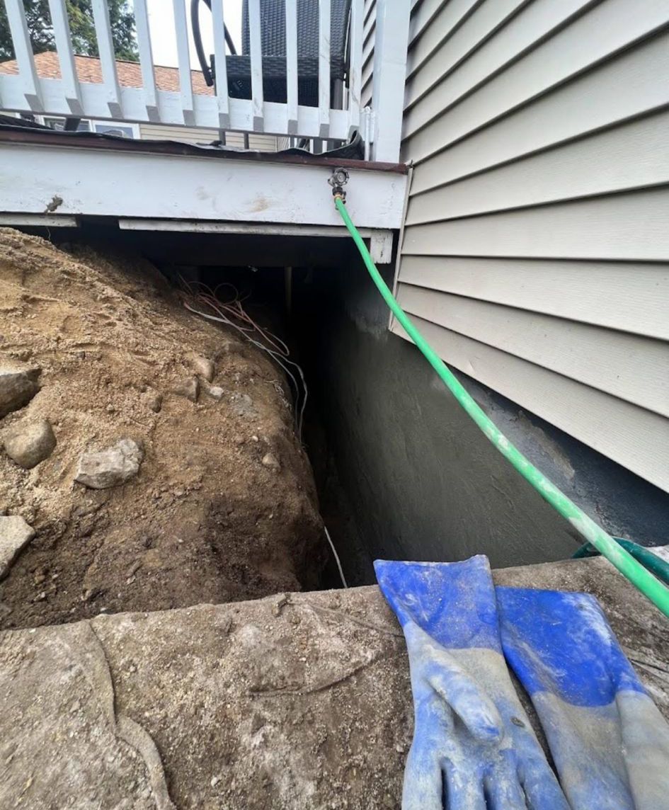 House foundation excavation with a green hose under a deck, dirt pile, and blue tarp in the foreground