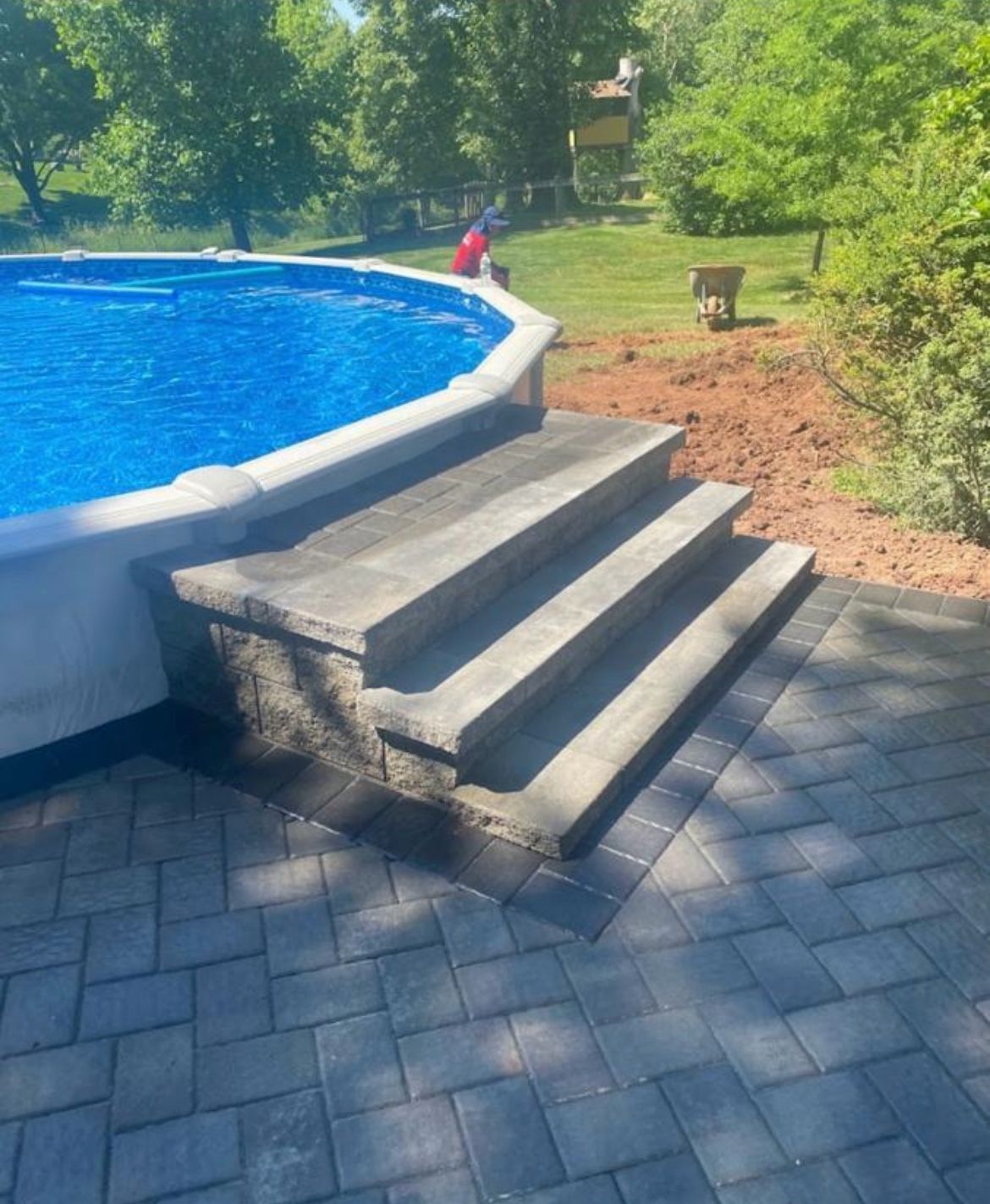 Backyard pool with stone steps, blue water, and paver patio beside green trees and shrubs