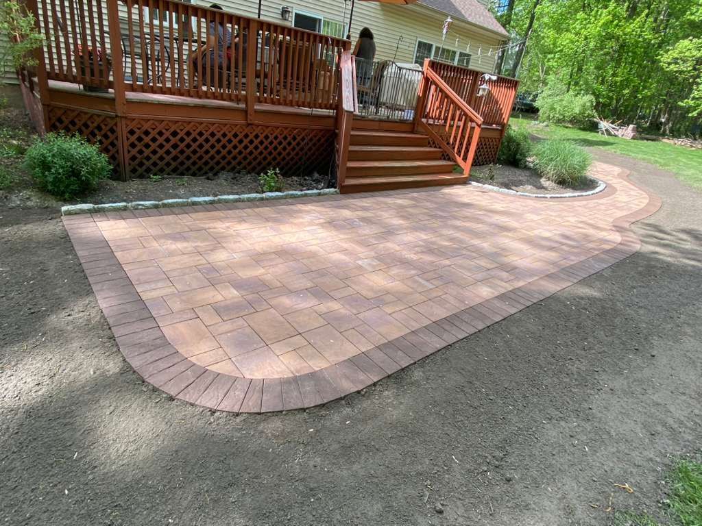 Brick paver patio and steps beside a wooden deck in a landscaped yard.