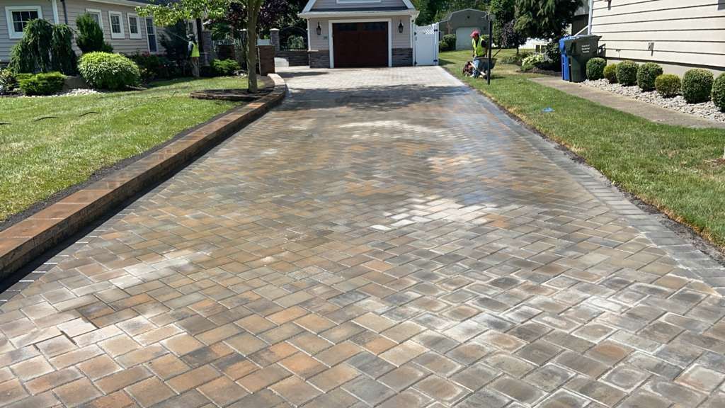 Brick driveway with white salt stains leading to a garage, bordered by green lawns and shrubs.