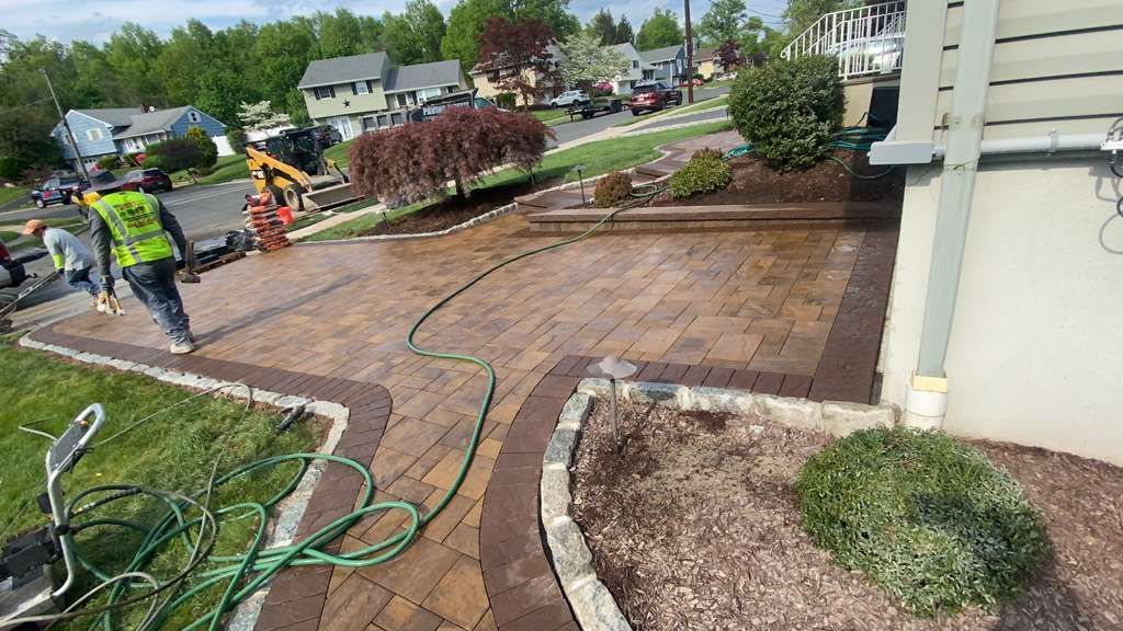 Workers installing pavers on a curved front walkway, with hoses and landscaping around a house.