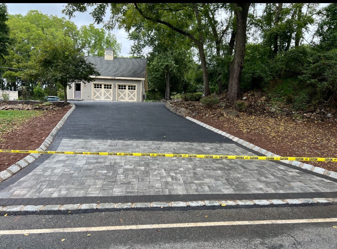 Freshly paved black driveway leading to a cream garage, bordered by trees and a yellow caution tape barrier