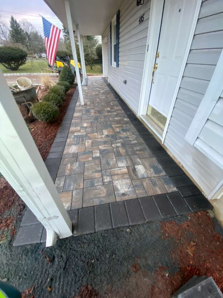 Covered front walkway with gray stone pavers beside a white house and an American flag planter box