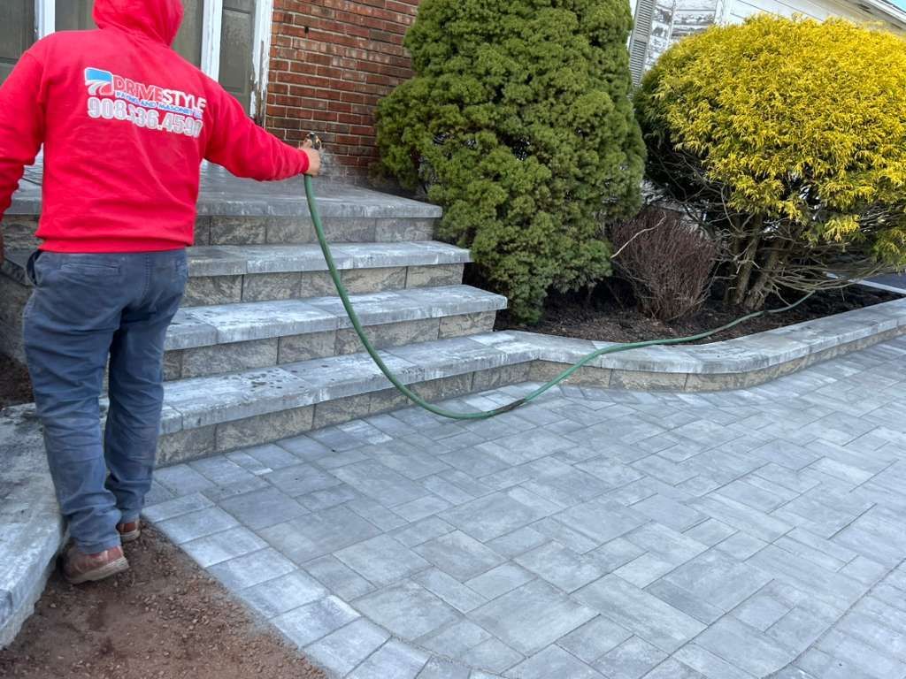 Person in a red hoodie using a hose to wash stone steps beside shrubs and a brick house