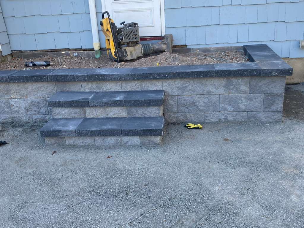Stone steps and retaining wall beside a house, with a yellow tool on the ledge and gravel foreground