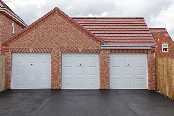 Three white garage doors on a red brick house with a dark driveway and wooden fence