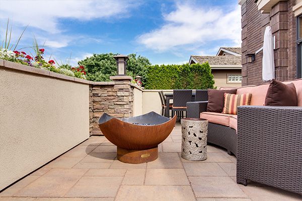 Patio with wicker sofa, round fire bowl, and stone planters under a blue sky