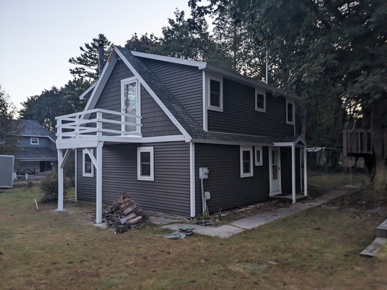 Two-story dark-gray house with white trim, balcony, and small windows, set in a grassy yard.