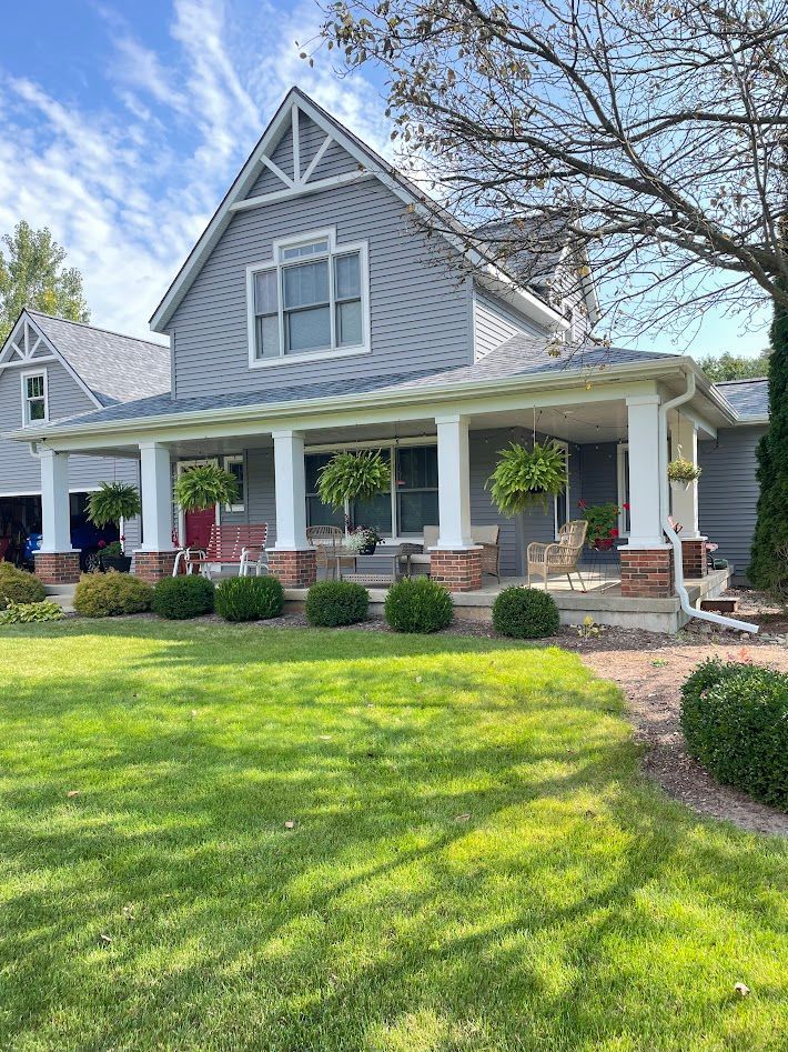 Gray house with a porch, green lawn, and hanging plants under a bright blue sky.
