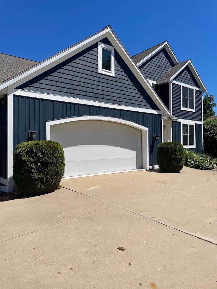Blue-sided house with white trim, a white garage door, and two small bushes in front. Clear, blue sky.