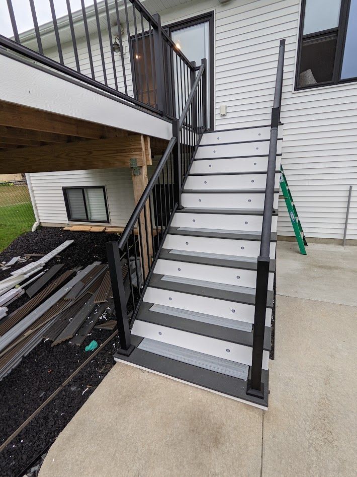 Staircase with black railing and composite steps in shades of gray leading from deck to concrete patio.