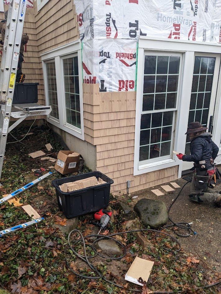 Exterior view of a home with a worker installing light brown shingles around a window.